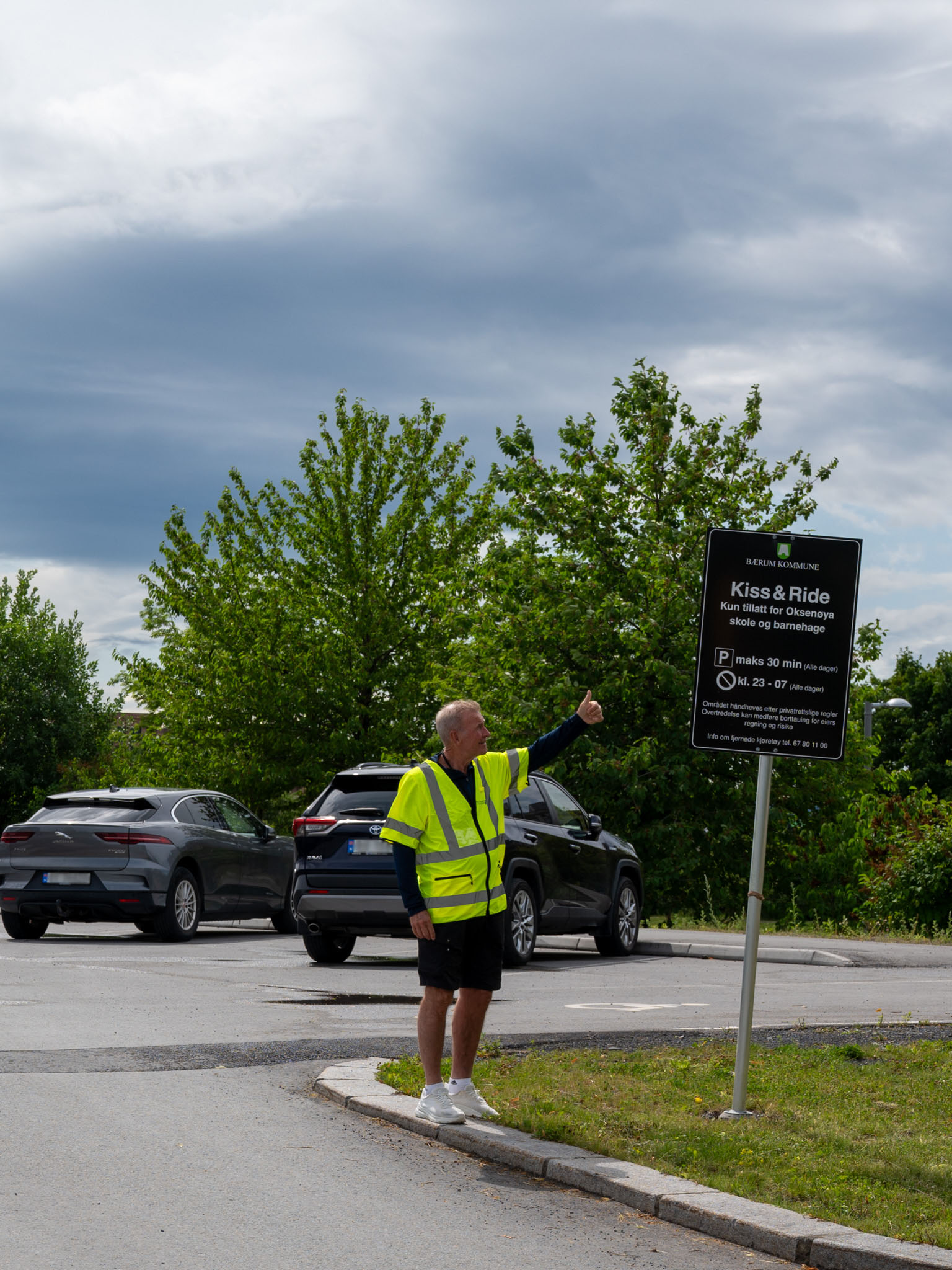 Dag Leween Stien, iført refleksvest, står ved et skilt i et parkeringsområde. Han peker på skiltet som har teksten: 'Kiss & Ride – Kort tid for av- og påstigning skole og barnehage.