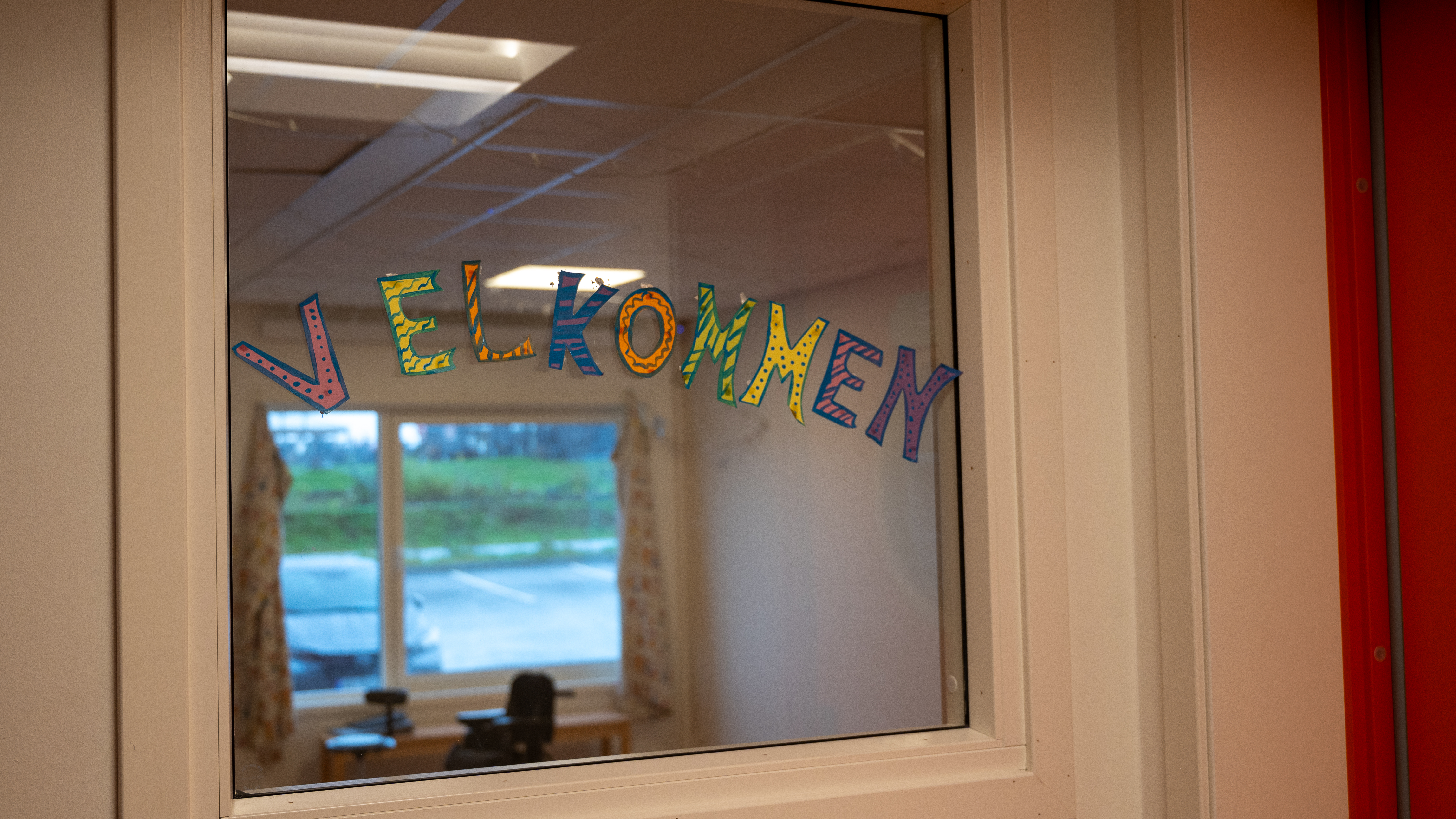 Et vindu med fargerike bokstaver som staver ordet 'Velkommen'. Vinduet gir utsikt til et rom med bord og stoler, mens et stort vindu i bakgrunnen. Bokstavene har ulike mønstre og farger.