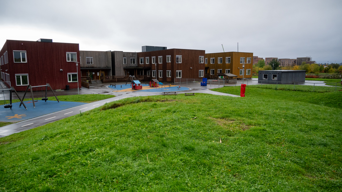 En stor barnehagebygning i flere farger, med to etasjer, står på en åpen gresslette. Foran bygningen er det en lekeplass med husker og små lekehus.
