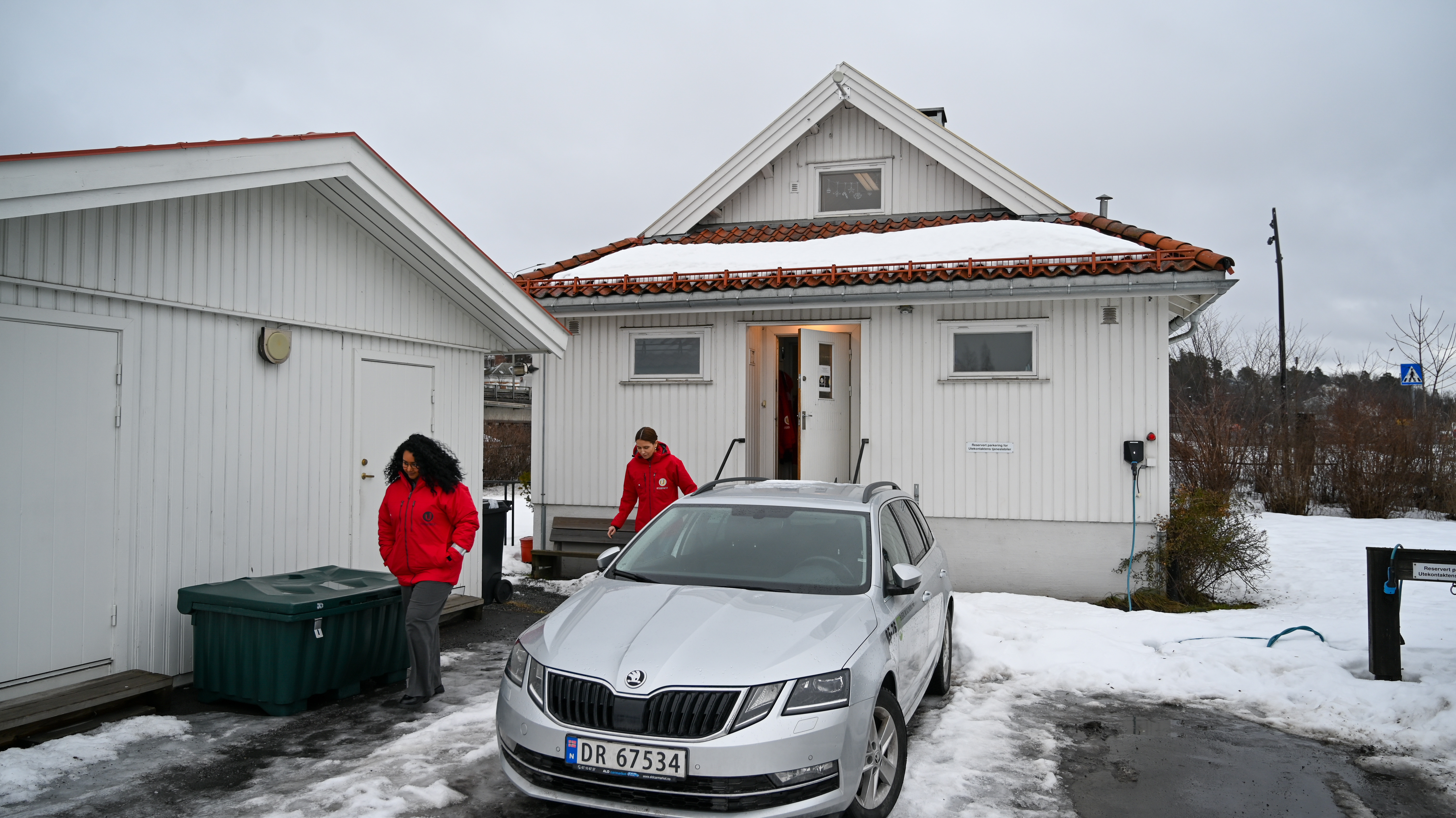Et hvitt trehus med rødt tak står i et vinterlig landskap. En sølvfarget bil er parkert foran, og to personer i røde jakker er på vei ut. Det er overskyet, og snø dekker bakken. Huset er lokalene til Utekontakten på Kadettangen i Sandvika.