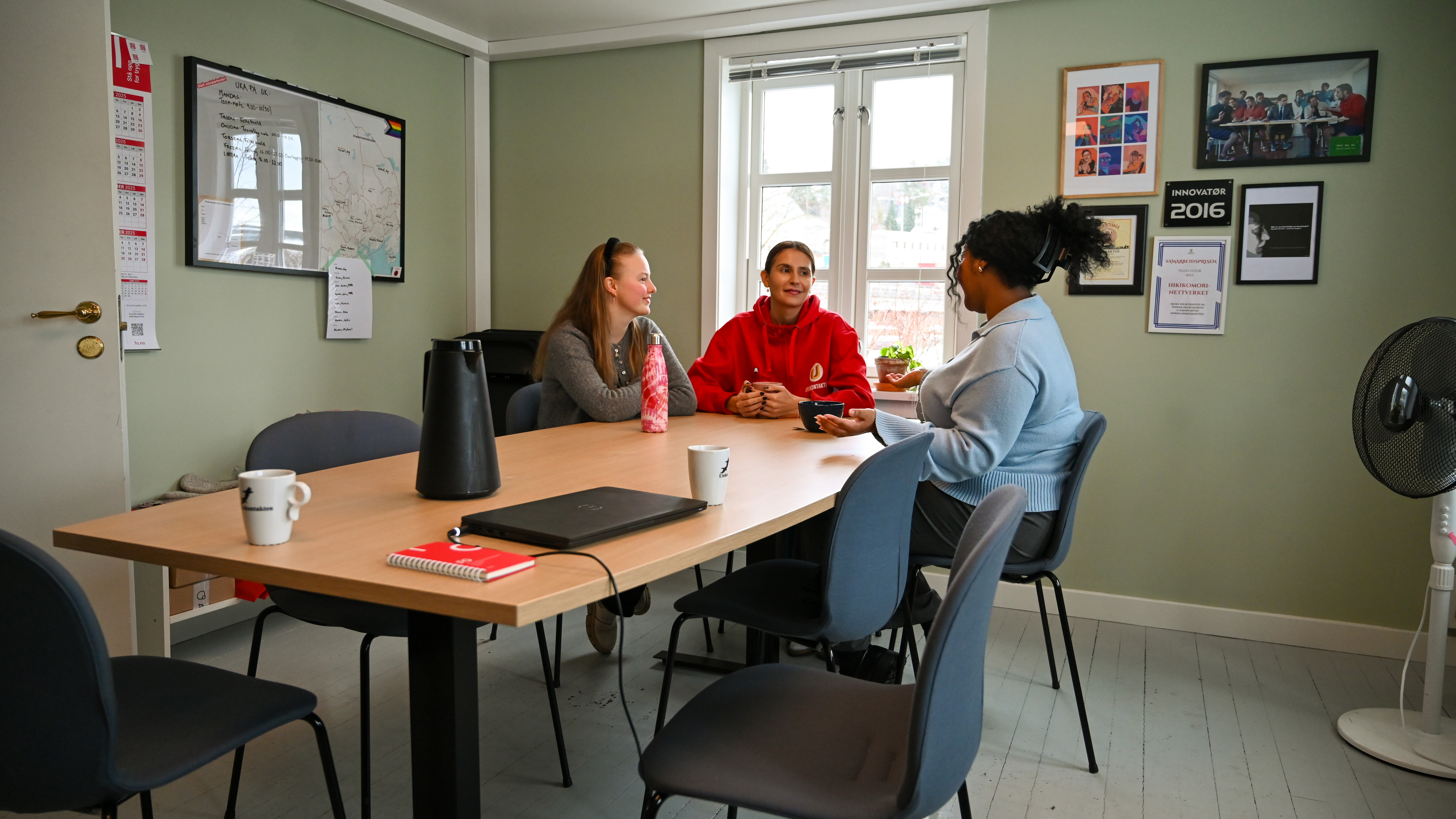 Tre kvinner sitter rundt et møtebord i et koselig og uformelt kontor- eller møterom. De er engasjert i samtale og ser ut til å ha en hyggelig diskusjon. Rommet har en stor tavle på veggen med en kartskisse, noen plakater og bilder på veggene, samt et vindu som slipper inn dagslys. Det er en avslappet atmosfære, og interiøret er enkelt og funksjonelt.